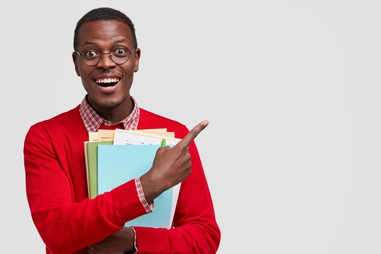 Positive college student has dark skin, carries folders and book, points with cheerful expression aside, has toothy smile, wears fashionable outfit, advertises something for studying isolated on white